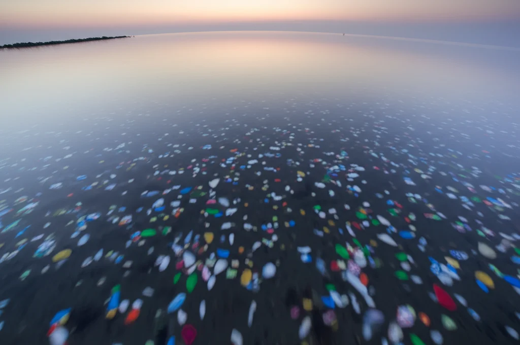Fotografia grandangolare della superficie dell'oceano durante un'alba nebbiosa, con l'acqua calma e riflettente. Sulla superficie sono sparsi numerosi piccoli detriti di plastica di vari colori e forme. Obiettivo grandangolare 18mm, lunga esposizione per rendere l'acqua setosa, messa a fuoco nitida su tutta la superficie, luce soffusa e colori pastello dell'alba.