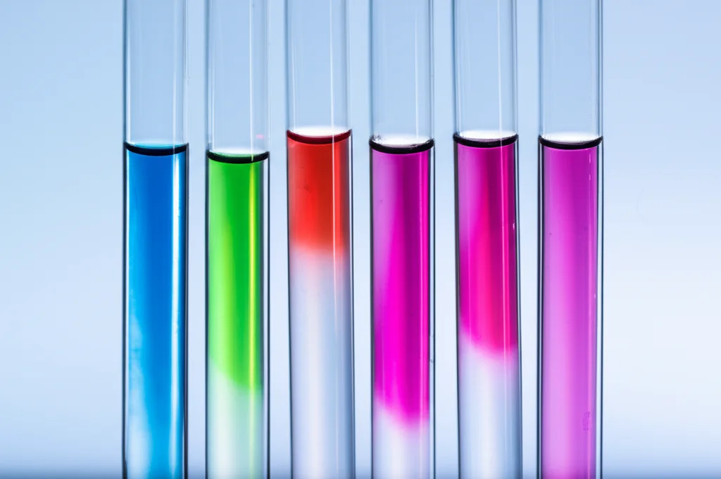 Fotografia di sette provette contenenti soluzioni acquose di diversi colori vivaci (blu, arancione, rosso, rosa, verde, viola) prima e dopo il trattamento con carbone attivo nero in polvere, mostrando la decolorazione. Still life, controlled lighting, high detail, macro lens 80mm.