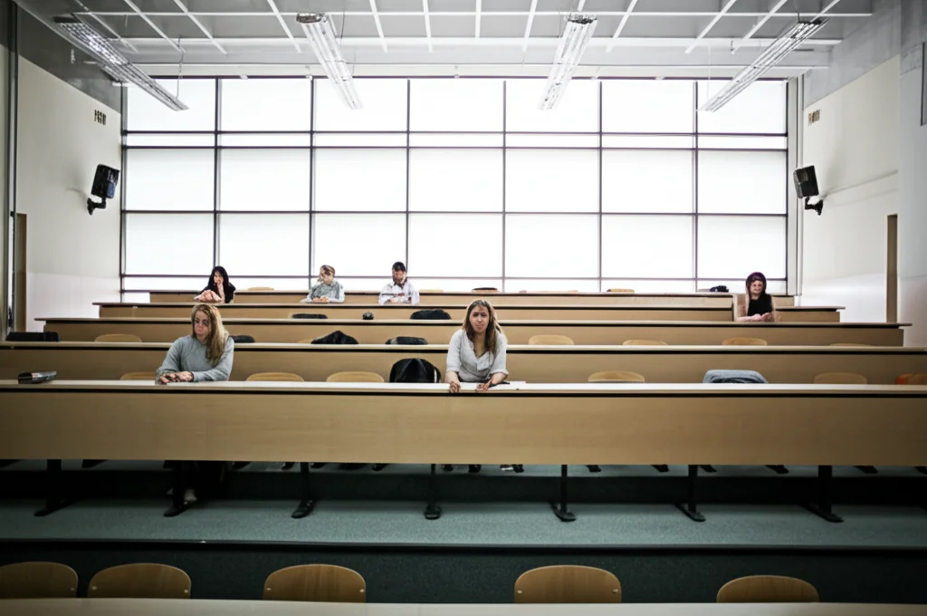 Fotografia grandangolare di un'aula universitaria di matematica o ingegneria, luminosa ma quasi vuota. Si vedono poche studentesse sparse tra molti posti vuoti, suggerendo il concetto di 'leaky pipeline'. Luce fredda proveniente da grandi finestre, obiettivo 20mm, messa a fuoco nitida sull'intera aula per dare un senso di vastità e vuoto.