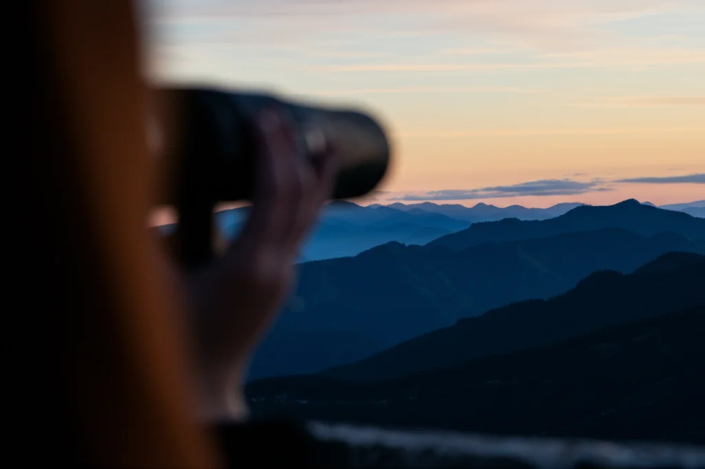 Un orizzonte lontano con montagne e cielo all'alba visto attraverso le lenti di un binocolo tenuto da mani leggermente sfocate in primo piano, paesaggio ampio e suggestivo, obiettivo grandangolare 15mm, messa a fuoco nitida sull'orizzonte per simboleggiare la ricerca futura, lunga esposizione per nuvole soffici.