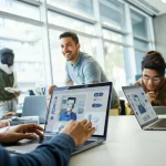 Un'aula universitaria luminosa e moderna con studenti diversi che collaborano attorno a un tavolo, alcuni usano laptop con interfacce AI stilizzate visibili sugli schermi, altri discutono animatamente. Focus su uno studente in primo piano che guarda sorridendo il suo schermo. Obiettivo prime 35mm, luce naturale diffusa da ampie finestre, profondità di campo media per mantenere il contesto visibile.