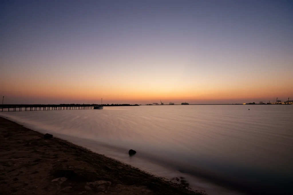 Veduta grandangolare del Lago Timsah, Egitto, al tramonto. L'acqua riflette i colori caldi del cielo. In lontananza si intravedono le sagome indistinte di navi o infrastrutture portuali, suggerendo l'attività umana nell'area. Obiettivo grandangolare 18mm, lunga esposizione per rendere l'acqua setosa, messa a fuoco nitida sull'orizzonte.