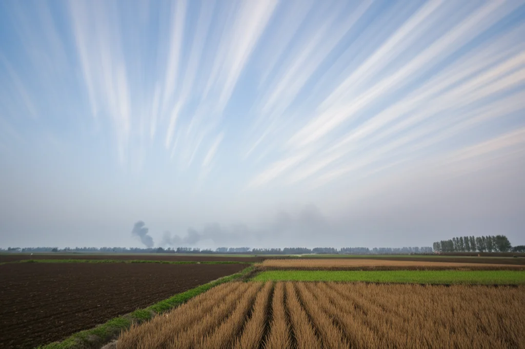 Fotografia grandangolare 15mm di campi agricoli cinesi con fumo da combustione di biomassa in lontananza sotto un cielo leggermente inquinato, luce del tardo pomeriggio, messa a fuoco nitida, lunga esposizione per nuvole mosse.
