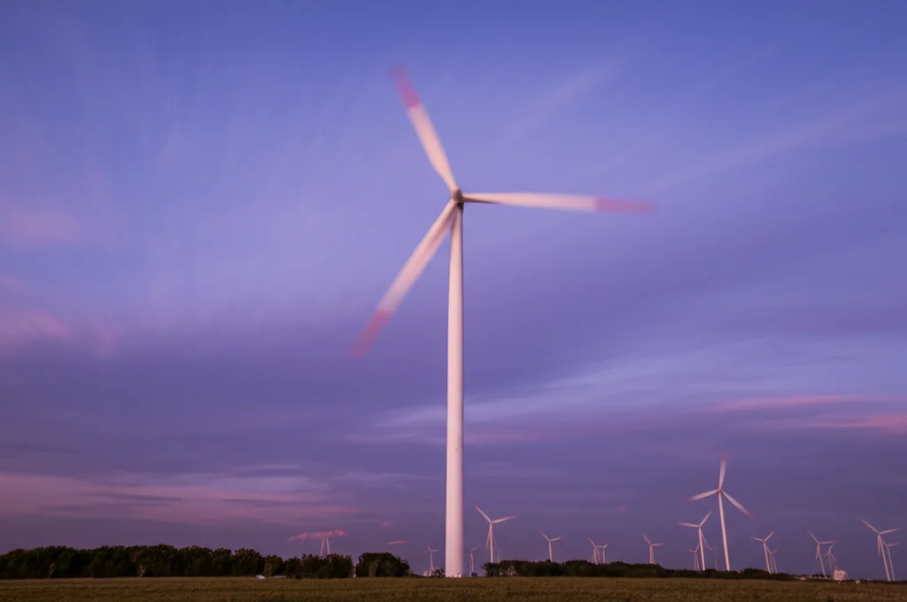 Turbina eolica maestosa in un parco eolico al tramonto, pale in leggero movimento rotatorio, fotografia grandangolare 15mm, lunga esposizione per creare scie soffuse delle nuvole nel cielo colorato, messa a fuoco nitida sulla struttura della turbina, paesaggio energetico.