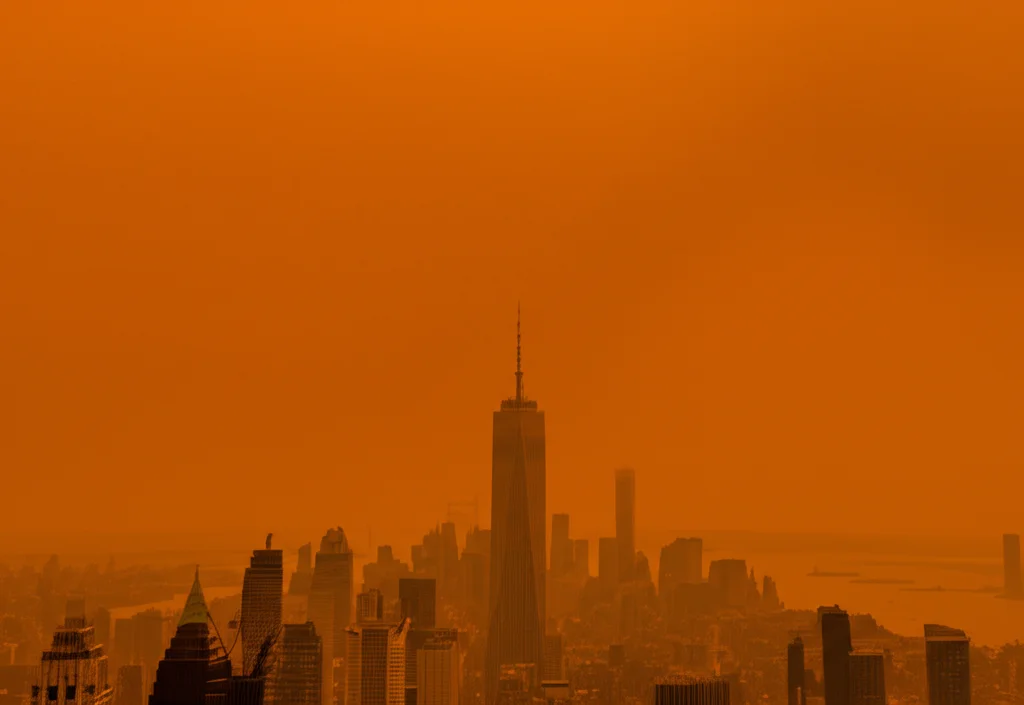Veduta aerea grandangolare (wide-angle, 15mm) del cielo sopra lo skyline di Manhattan tinto di un arancione intenso a causa del fumo degli incendi canadesi, con focus nitido (sharp focus) sulle particelle sospese nell'aria, lunga esposizione per accentuare l'effetto foschia.