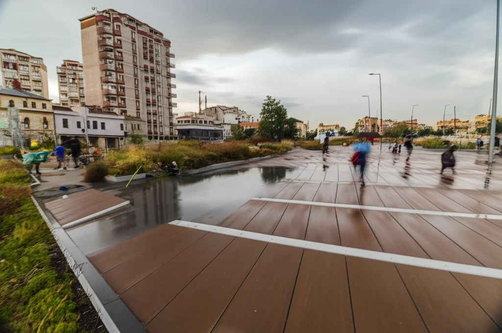 Fotografia grandangolare di un paesaggio urbano resiliente dopo un evento climatico (es. pioggia intensa). Si vedono infrastrutture verdi (tetti giardino, aree permeabili) e persone che collaborano per ripulire. Obiettivo grandangolare 18mm, lunga esposizione per mostrare movimento residuo dell'acqua o delle nuvole, messa a fuoco nitida su tutto il panorama, luce del tardo pomeriggio.