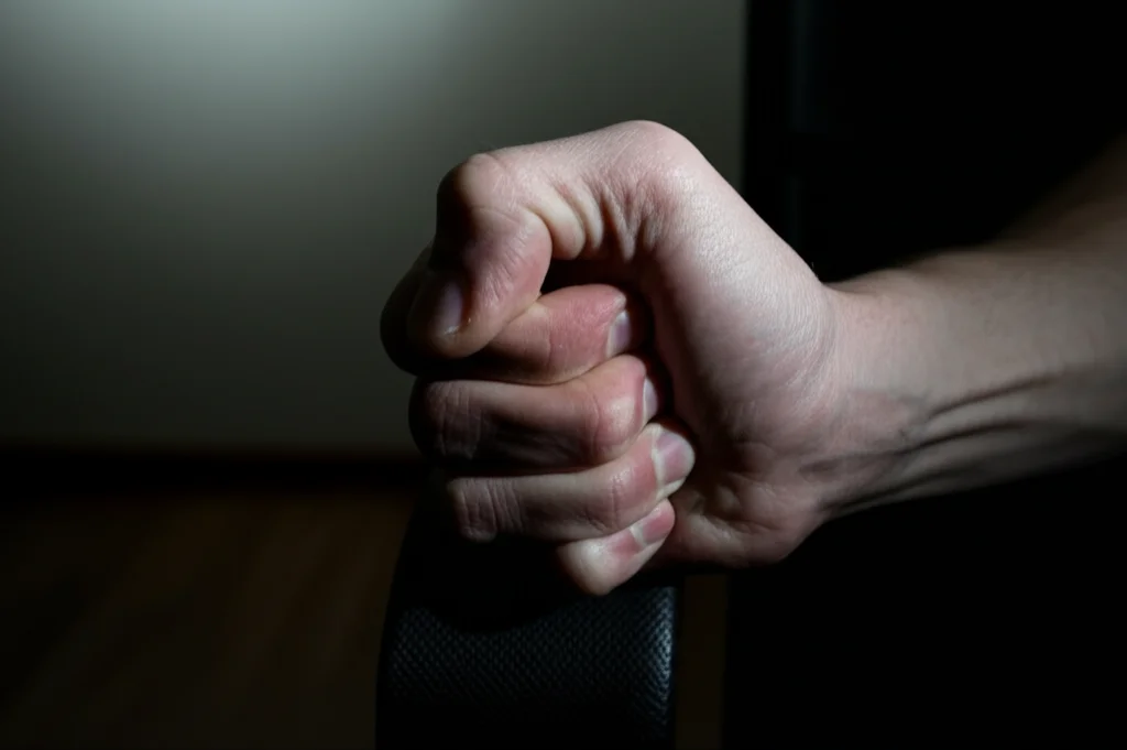 Fotografia macro, 90mm, di una mano che stringe forte il bracciolo di una sedia in una stanza spoglia, illuminazione controllata che crea ombre profonde, alta definizione sui dettagli delle nocche bianche per esprimere tensione emotiva, colpa e conflitto interiore.