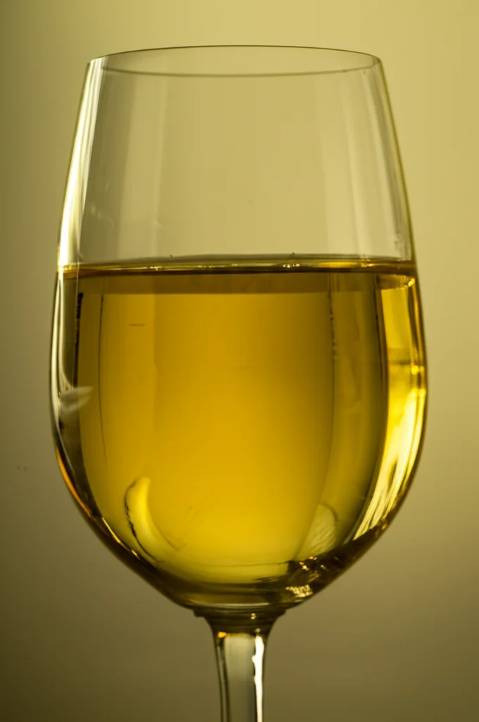 Primo piano di un bicchiere di vino Chardonnay dal colore giallo dorato intenso, controluce per evidenziare la brillantezza e la saturazione del colore, obiettivo prime 50mm, profondità di campo ridotta.