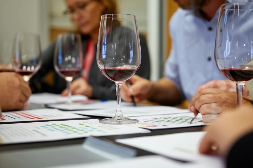 Fotografia di un gruppo di persone durante una degustazione professionale di vino in un laboratorio sensoriale, focus sui bicchieri e sulle schede di valutazione, obiettivo zoom 70mm, luce naturale soffusa.