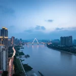 Fotografia paesaggistica grandangolare (obiettivo 10-24mm) dello skyline di Chongqing o delle sue montagne circostanti durante la transizione tra la notte e le prime luci dell'alba. Il cielo mostra colori pre-alba con nuvole basse o foschia persistente nelle valli, suggerendo l'umidità e il fenomeno delle piogge notturne. Possibile lunga esposizione per catturare le luci della città e rendere l'acqua del fiume Yangtze setosa. Messa a fuoco nitida sul panorama.