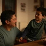Ritratto fotografico di un genitore Li sorridente che interagisce con il proprio figlio in età scolare primaria in un ambiente domestico caldo e accogliente, obiettivo 50mm, luce naturale morbida, profondità di campo che isola i soggetti.