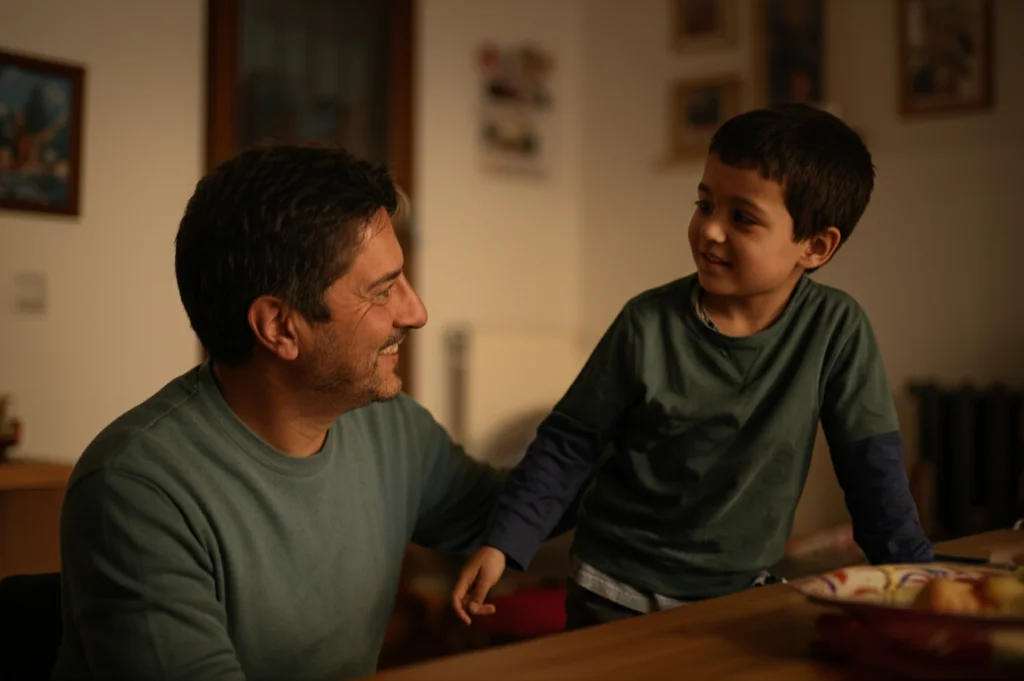 Ritratto fotografico di un genitore Li sorridente che interagisce con il proprio figlio in età scolare primaria in un ambiente domestico caldo e accogliente, obiettivo 50mm, luce naturale morbida, profondità di campo che isola i soggetti.