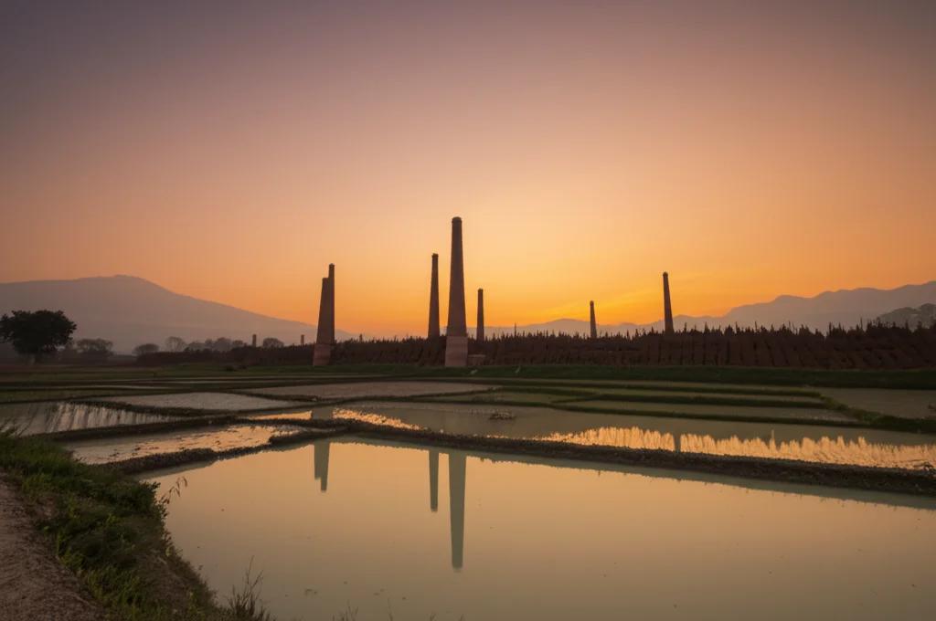 Fotografia grandangolare di un paesaggio rurale al tramonto in Nepal, si vedono in lontananza ciminiere di fornaci di mattoni. Obiettivo grandangolare 18mm, lunga esposizione per catturare i colori caldi del cielo e rendere l'acqua di un eventuale fiume liscia come seta, messa a fuoco nitida sull'intero paesaggio per dare un senso di vastità e contesto.