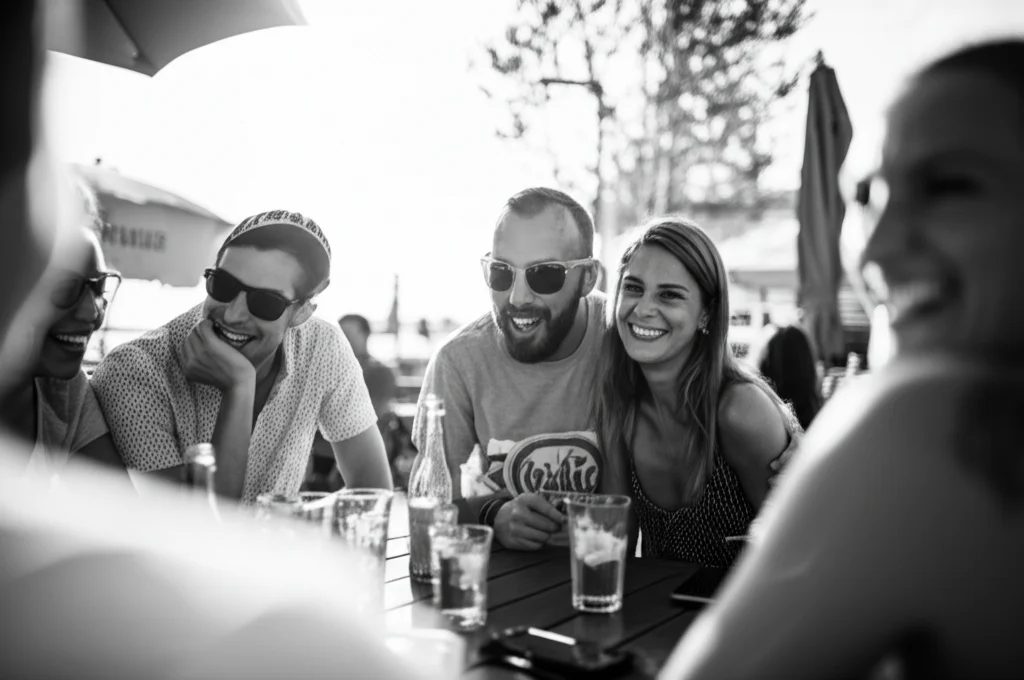 Ritratto di gruppo di amici che ridono attorno a un tavolo all'aperto in un pomeriggio soleggiato, obiettivo 35mm, luce naturale, profondità di campo media per mantenere a fuoco il gruppo ma sfocare leggermente lo sfondo, bianco e nero.