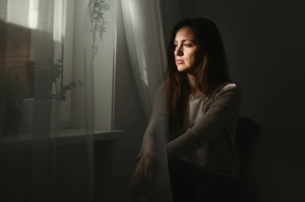 Una donna seduta vicino a una finestra, guarda fuori pensierosa ma non triste, con una luce soffusa che entra. Fotografia ritratto, obiettivo 50mm, toni caldi e leggermente desaturati, profondità di campo per isolare la figura.