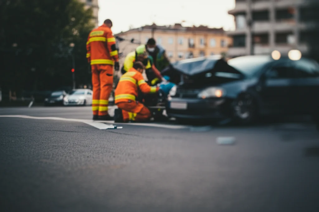 Scena di un incidente stradale urbano post-pandemia, con soccorritori che assistono un ferito vicino a un'auto danneggiata. Obiettivo 35mm, luce del tardo pomeriggio, profondità di campo ridotta per focalizzare sull'emergenza.