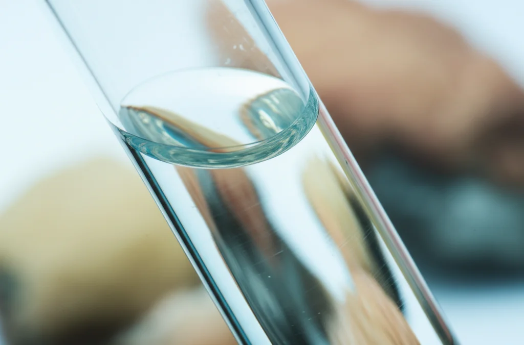 Fotografia macro di un campione d'acqua limpida in una provetta da laboratorio, con rocce silicate e solfatiche sfocate sullo sfondo a rappresentare l'interazione acqua-roccia, illuminazione controllata per dettagli precisi, obiettivo macro 100mm, alta definizione.