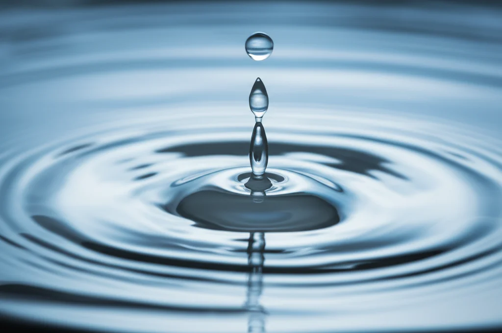 Primo piano macro di una goccia d'acqua cristallina che cade in uno specchio d'acqua calma, creando piccole increspature concentriche. Obiettivo macro 100mm, alta definizione, illuminazione laterale controllata per enfatizzare la trasparenza e i riflessi.