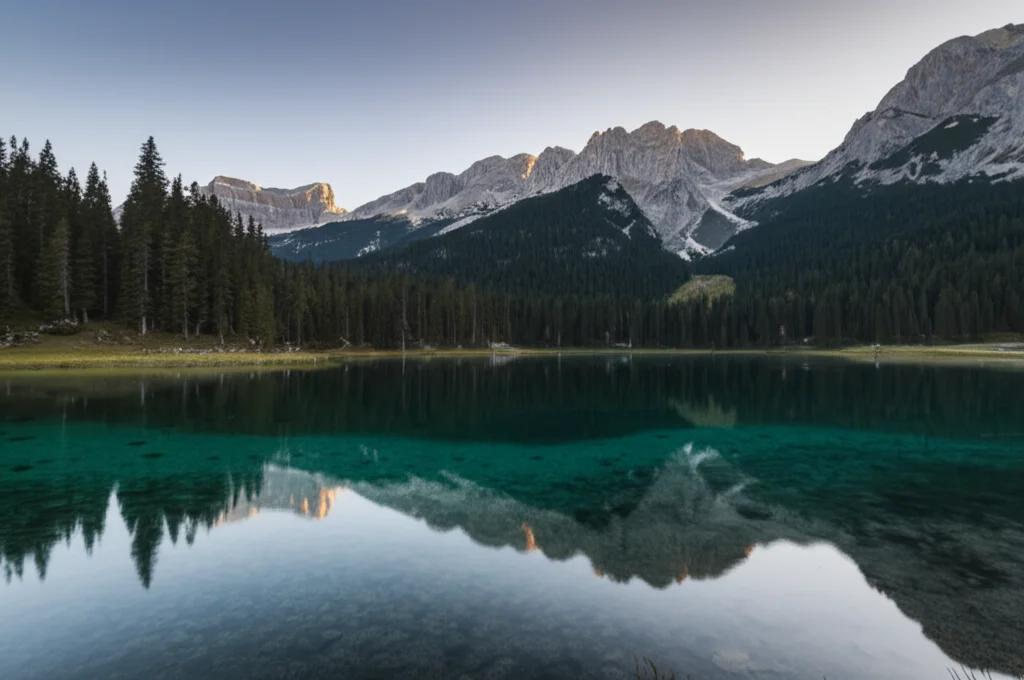 Fotografia paesaggistica grandangolare di un lago alpino cristallino al mattino presto, con le montagne che si riflettono perfettamente sull'acqua liscia. Obiettivo grandangolare 14mm, lunga esposizione per effetto specchio, messa a fuoco nitida su tutto il paesaggio, colori naturali e vibranti.