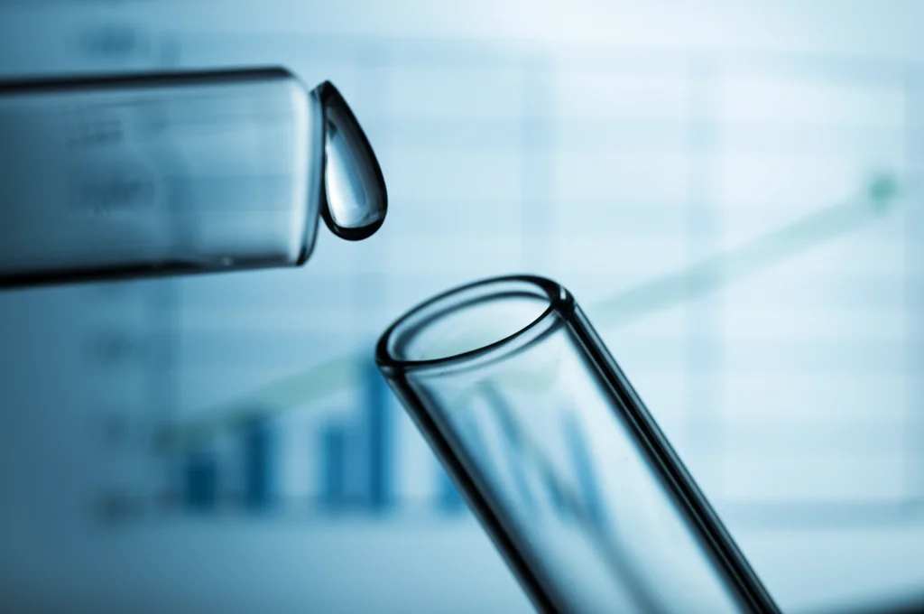 Primo piano macro di una goccia di saliva su una provetta di vetro da laboratorio, illuminazione controllata per evidenziare la trasparenza. Obiettivo macro 100mm, alta definizione, messa a fuoco precisa sulla goccia. Sullo sfondo sfocato, grafici stilizzati che mostrano un aumento degli antiossidanti e una diminuzione dei marcatori di stress ossidativo.