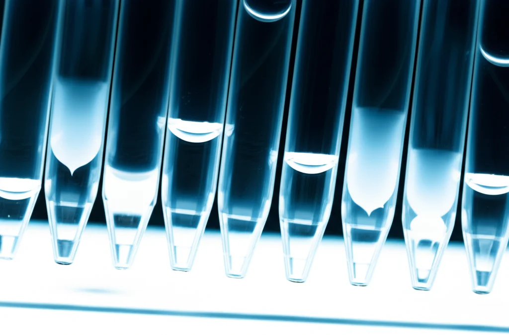 Fotografia macro che mostra una serie di piccole provette Eppendorf contenenti volumi decrescenti (da sinistra a destra) di un residuo polimerico secco sul fondo. Obiettivo macro 60mm, illuminazione da laboratorio, messa a fuoco precisa sui residui, simulando esperimenti a volume ridotto.