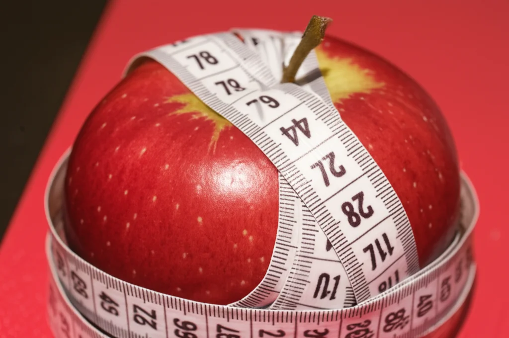 Fotografia macro di un metro a nastro avvolto intorno a una mela rossa matura, illuminazione controllata e precisa, alta definizione dei dettagli sulla buccia della mela e sul nastro, obiettivo macro 100mm, a simboleggiare la misurazione della salute e del rischio cardiometabolico.