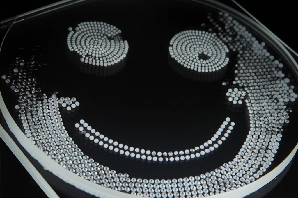 Macro fotografia, lente 85mm, sequenza di immagini che mostrano la pulizia progressiva di microsfere seguendo un pattern a forma di faccina sorridente su una lastrina di vetro, evidenziando la rimozione selettiva guidata dall'AI, illuminazione controllata. High detail, precise focusing.