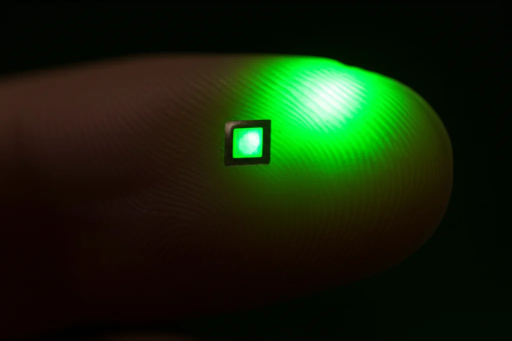 Fotografia macro di un sensore PPG a luce verde posizionato su un dito indice, messa a fuoco precisa sul sensore e sulla pelle, illuminazione controllata per evidenziare i dettagli, obiettivo macro 90mm.