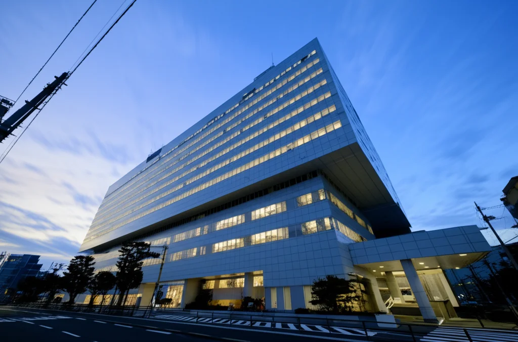 Scatto grandangolare di un moderno edificio ospedaliero giapponese visto dall'esterno durante l'ora blu, poco dopo il tramonto. Le luci sono accese all'interno delle finestre. Il cielo ha sfumature di blu e arancione. Lunga esposizione utilizzata per creare un effetto liscio sulle poche nuvole presenti. Obiettivo grandangolare 14mm, messa a fuoco nitida su tutto l'edificio.