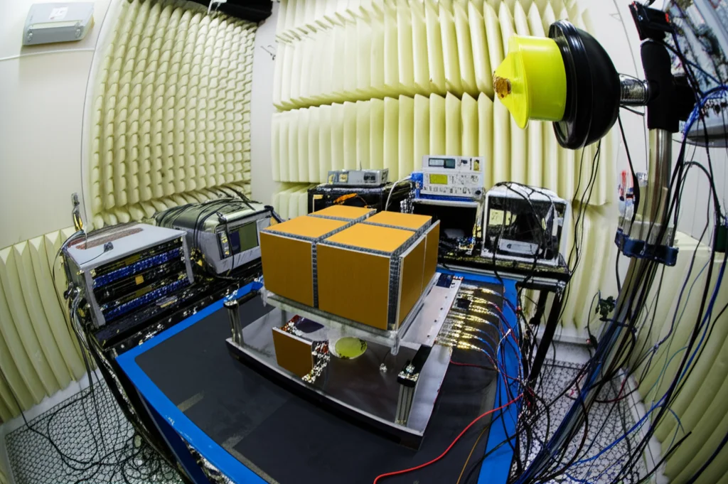 Immagine dell'interno di una camera anecoica durante un esperimento di energy harvesting. Un array di metamateriali 3x3 è montato su un supporto, puntato verso un'antenna trasmittente. Strumenti di misura visibili sullo sfondo. Focus nitido sull'array, wide-angle 20mm, illuminazione tecnica.