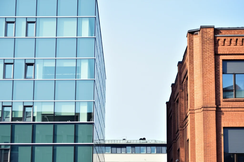 Scatto grandangolare, obiettivo 15mm, che mostra due edifici aziendali distinti affiancati. Uno moderno in vetro (simbolo tech?), l'altro tradizionale in mattoni (simbolo manifatturiero?), a simboleggiare la fusione di industrie diverse. Messa a fuoco nitida, luce diurna.