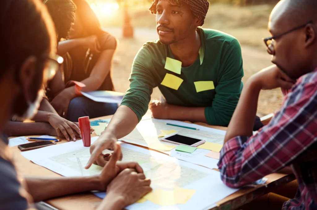 Fotografia a colori vivaci, obiettivo 50mm, che ritrae un gruppo diversificato di persone (scienziati, membri della comunità, giovani) seduti in cerchio all'aperto durante un workshop partecipativo, discutono animatamente indicando mappe e grafici sparsi su un tavolo rustico, luce naturale pomeridiana calda.