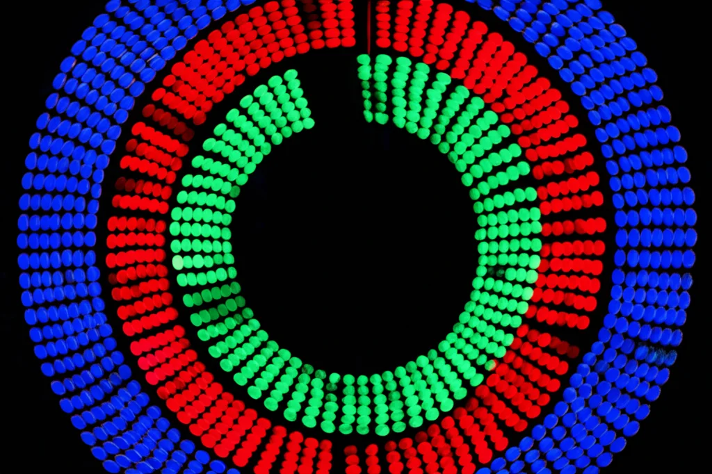 Immagine fotorealistica di un display sperimentale dell'Esperimento 2: anelli concentrici di punti luminosi (verde al centro, rosso interno, blu esterno) su sfondo nero, catturati in movimento. Teleobiettivo zoom 150mm, fast shutter speed per congelare parzialmente il movimento ma suggerirne la dinamica, action tracking.