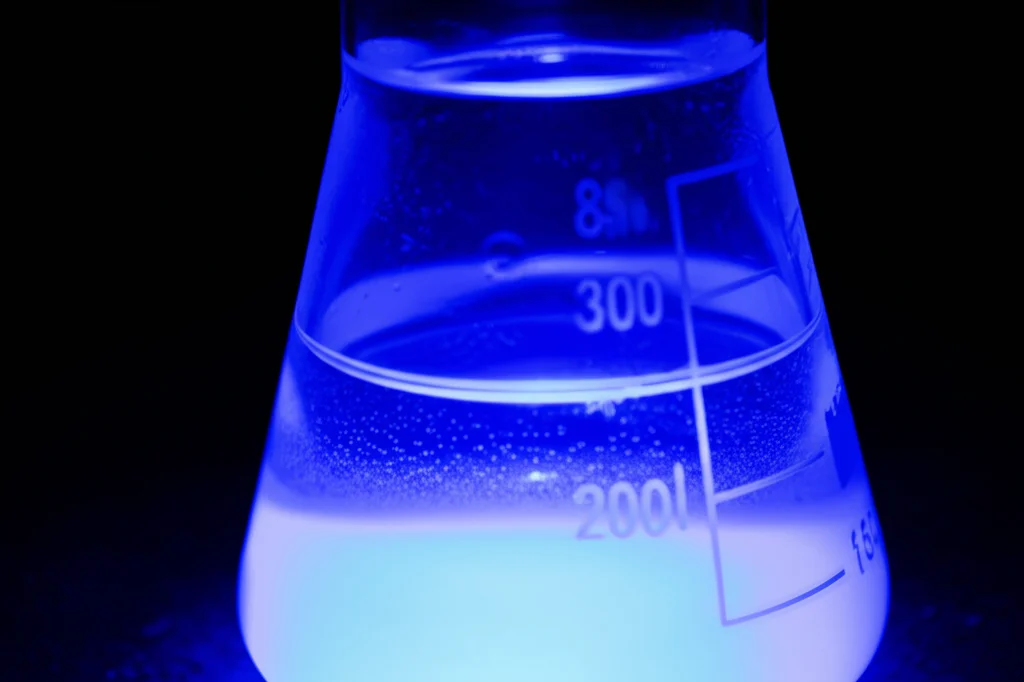 Fotografia di un setup di laboratorio per la fotocatalisi: un becher contenente acqua blu (simulando il Blu di Metilene) con nanoparticelle di ZnO in sospensione, illuminato dall'alto da una lampada UV. L'acqua mostra un visibile schiarimento vicino alla superficie. Obiettivo prime 35mm, profondità di campo che isola il becher, sfondo scuro per enfatizzare la luce UV.