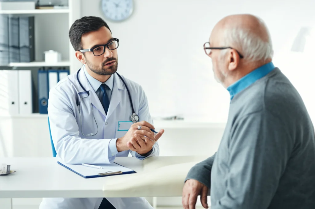 Fotografia di un medico cardiologo che spiega dei risultati a un paziente anziano in uno studio medico luminoso. L'atmosfera è seria ma empatica, focalizzata sulla comunicazione e la cura a lungo termine. Obiettivo prime 50mm, profondità di campo che isola medico e paziente, luce naturale.