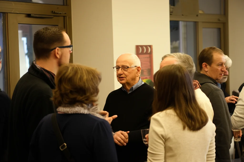 Gruppo eterogeneo di persone di età diverse (adolescenti, giovani adulti, mezza età, anziani) che conversano animatamente in un ambiente informale e luminoso, teleobiettivo zoom 150mm per catturare interazioni naturali, fast shutter speed.