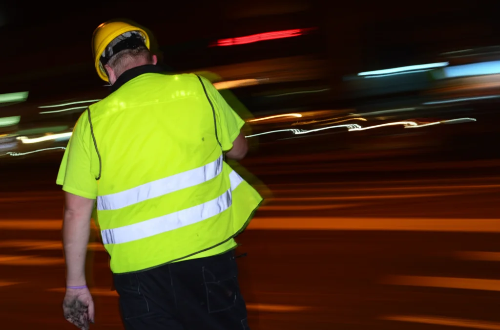 Fotografia in movimento di un operaio stradale di notte che indossa un gilet ad alta visibilità in tessuto misto poliestere/cotone tinto con colorante fluorescente giallo. Il gilet brilla sotto le luci artificiali mentre l'operaio lavora. Teleobiettivo zoom 150mm, fast shutter speed per congelare il movimento, leggero motion blur sullo sfondo stradale.