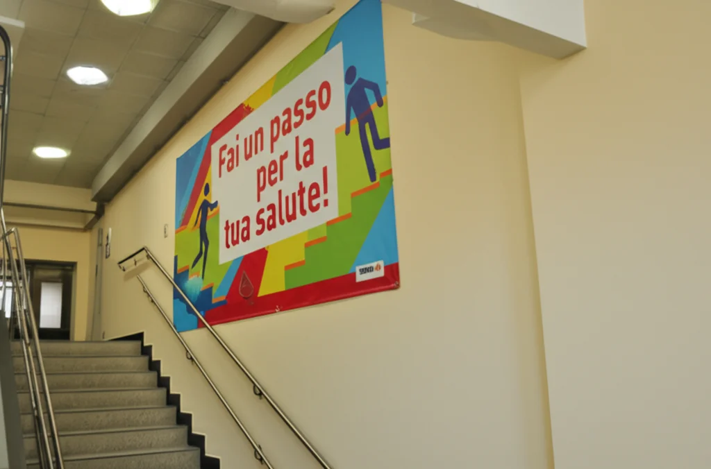 Dettaglio di un grande banner colorato appeso al muro accanto a una rampa di scale in un ambiente universitario. Il banner mostra slogan motivazionali come 'Fai un passo per la tua salute!' e icone stilizzate di persone che salgono le scale. Obiettivo macro 100mm, alta definizione dei dettagli del tessuto e della stampa, illuminazione laterale controllata per dare tridimensionalità.