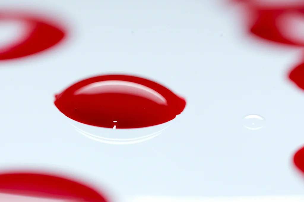 Primo piano macro di una goccia di sangue su una piastra da laboratorio, illuminazione controllata e precisa, alta definizione dei componenti sanguigni, lente macro 100mm, focus selettivo.