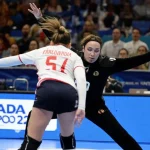 Foto di pallamano femminile ad alta azione, teleobiettivo 300mm, velocità otturatore elevata, tracciamento del movimento, una giocatrice segna un gol superando il portiere durante una partita intensa del Campionato Europeo 2022, con tifosi sfocati sullo sfondo.