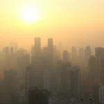 Skyline di una città del sud-est asiatico parzialmente oscurato da una foschia da inquinamento atmosferico, con il sole che filtra debolmente attraverso le particelle sospese. Fotografia grandangolare con obiettivo da 24mm, lunga esposizione per accentuare l'effetto della foschia e rendere l'acqua liscia se presente, focus nitido sugli edifici in primo piano per contrasto.