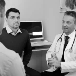 Un medico e un paziente seduti uno di fronte all'altro in un moderno studio medico, impegnati in una conversazione attenta. Il medico guarda il paziente con empatia, mentre il paziente sembra esprimere le proprie preoccupazioni. L'immagine trasmette fiducia e professionalità. Prime lens, 35mm, black and white film, depth of field per un look classico e intenso.