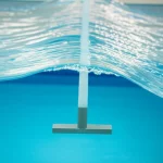 Fotografia realistica di un'onda regolare che si propaga su una barra sommersa visibile sotto l'acqua limpida in un canale ondoso sperimentale. Obiettivo 35mm, luce controllata da laboratorio, messa a fuoco nitida sull'onda, leggera turbolenza visibile dopo la barra. Profondità di campo che mantiene a fuoco sia l'onda che la barra.