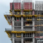 Immagine di un cantiere di un grattacielo con casseforme in alluminio e tunnel in primo piano, telephoto zoom, 100mm, action or movement tracking, operai al lavoro, cielo parzialmente nuvoloso.