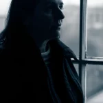 Un ritratto fotografico toccante, obiettivo 35mm, di una persona adulta che guarda fuori da una finestra in una giornata nuvolosa, il suo volto parzialmente in ombra riflette una profonda introspezione, duotone blu e grigio per accentuare la malinconia, profondità di campo che sfoca lo sfondo, simboleggiando l'impatto duraturo delle esperienze infantili avverse sulla vita adulta.
