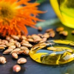 Un'immagine macro ad alta definizione di luminosi semi di cartamo e gocce dorate di olio di cartamo su una superficie scura e testurizzata, con un fiore di cartamo arancione brillante parzialmente a fuoco sullo sfondo. Lente macro 105mm, illuminazione controllata per esaltare texture e colori, focus preciso sui semi e sull'olio.