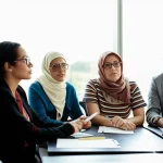 Immagine fotorealistica che simboleggia l'educazione sanitaria inclusiva: un gruppo diversificato di persone, alcune con ausili visivi o uditivi, partecipa attivamente a una sessione formativa in un ambiente moderno e luminoso. Obiettivo da 35mm, luce naturale diffusa, espressioni concentrate e positive.