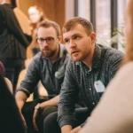 A diverse group of adults, some appearing neurotypical, some autistic, engaged in conversation, with subtle visual cues suggesting different communication styles. Shot with a 35mm prime lens, capturing depth of field, slightly desaturated colors for a thoughtful mood.