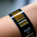 Primo piano di un sensore indossabile futuristico per l'analisi dei gas cutanei, appoggiato delicatamente su un avambraccio, con dettagli high-tech visibili. Obiettivo macro 90mm, illuminazione da studio controllata, alta definizione dei materiali, sfondo leggermente sfocato per concentrare l'attenzione sul dispositivo.