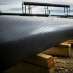 Confronto visivo tra un moderno tubo strutturale in CFRP nero e un tubo tradizionale in acciaio arrugginito, utilizzati come pali di fondazione in un cantiere. Obiettivo prime 35mm, profondità di campo, luce naturale drammatica che evidenzia la modernità del CFRP e il degrado dell'acciaio.